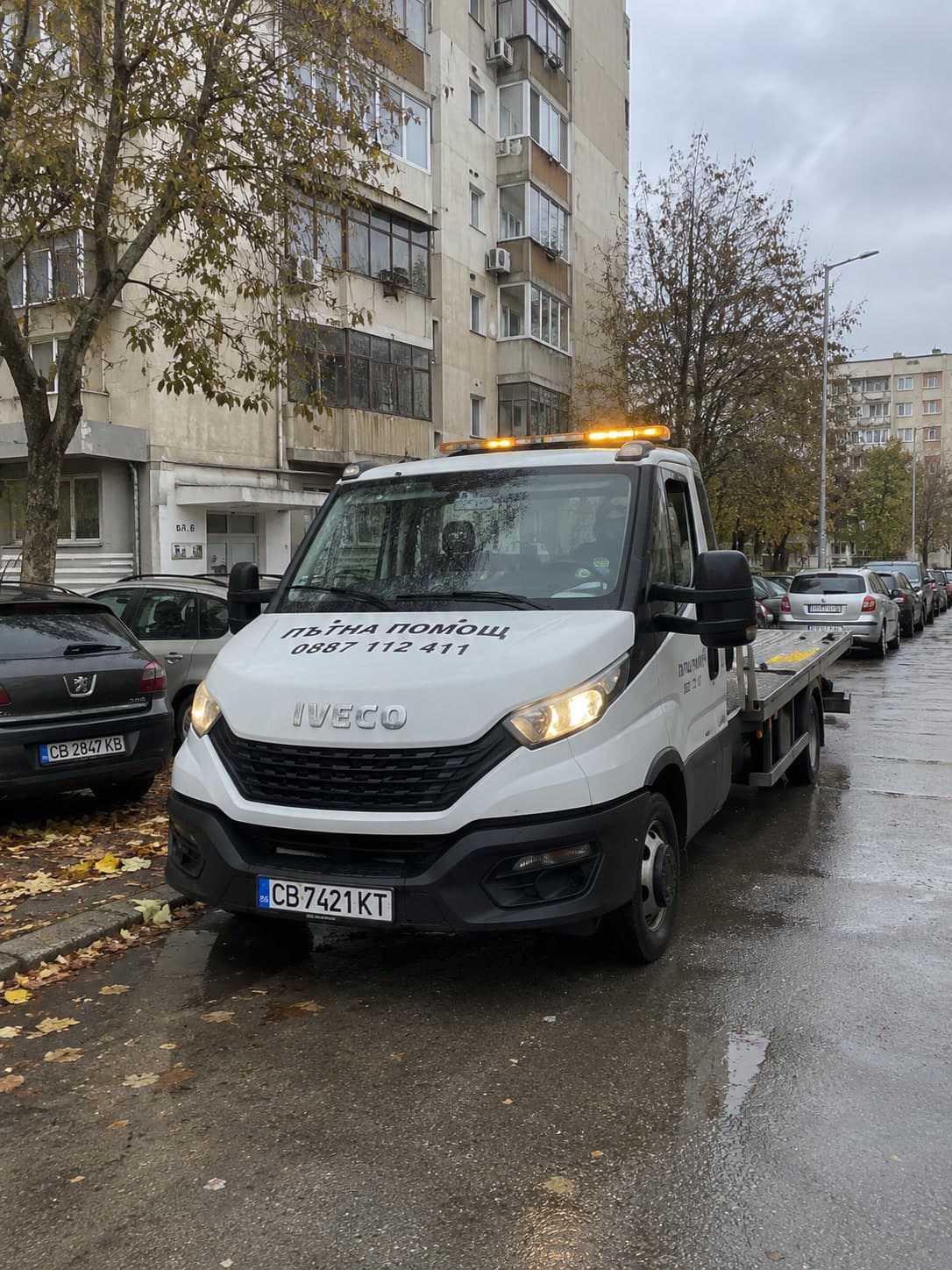 Платформата спира пред блок в дъждовно време, със светещи аварийни светлини.