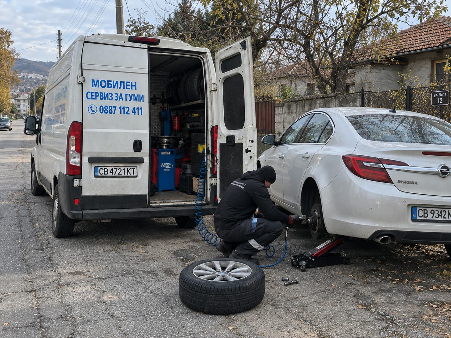 Работа по гума на бял автомобил пред къща, с крик и пневматичен гайковерт.